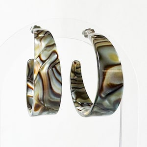 May include: A pair of hoop earrings with a marbled pattern in shades of brown, tan, and gray. The earrings have a rounded, squared-off shape and are displayed on a clear stand against a white background. The earrings are a fashion accessory.