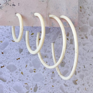 May include: Set of four white hoop earrings of varying sizes. The earrings are made of a smooth, white material and are displayed on a pink background.