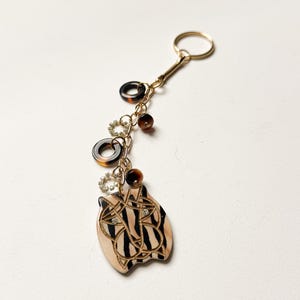 May include: A gold-toned keychain with a decorative charm. The charm features a light tan oval shape with a black and gold design. The keychain also includes brown and gold accents, and a gold ring.