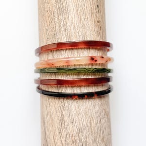 Puede incluir: Colección de seis pulseras finas en varios colores, incluyendo rojo, verde y carey, apiladas en una clavija de madera. Las pulseras tienen un acabado liso y pulido y están hechas de un material translúcido.