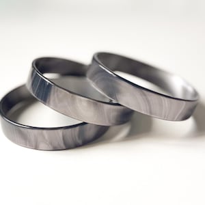 Può includere: Tre braccialetti grigi con effetto marmorizzato sono impilati su una superficie bianca. I braccialetti hanno una finitura liscia e lucida e un motivo sottile e vorticoso. I braccialetti sono un accessorio semplice ed elegante.