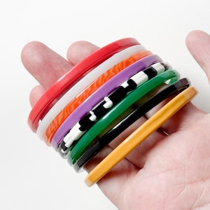 Op de afbeelding: Een verzameling kleurrijke armbanden in rood, wit, oranje, paars, zwart en wit, groen, zwart en geel. De armbanden zijn op elkaar gestapeld en tonen een verscheidenheid aan levendige tinten en patronen. De armbanden worden in een hand gehouden.
