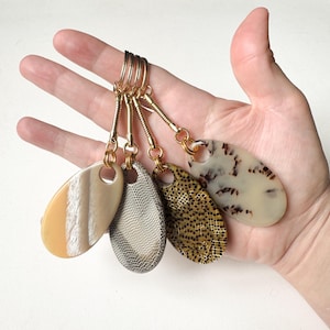 Könnte beinhalten: Eine Hand, die fünf Schlüsselanhänger hält. Jeder Schlüsselanhänger hat einen goldfarbenen Ring und eine Kette, die mit einem ovalen Anhänger mit einzigartigen Mustern verbunden sind, darunter Streifen, Mesh und ein gesprenkeltes Design.