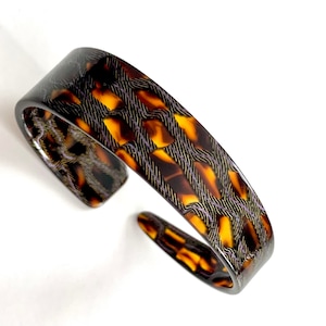 May include: A brown and black patterned bangle bracelet. The pattern is a geometric design with a woven look.