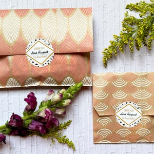 May include: Two small gift bags with a pink and gold geometric pattern. Each bag has a round sticker with black and white polka dots and the text "THANK YOU Lisa August For Your Order!"