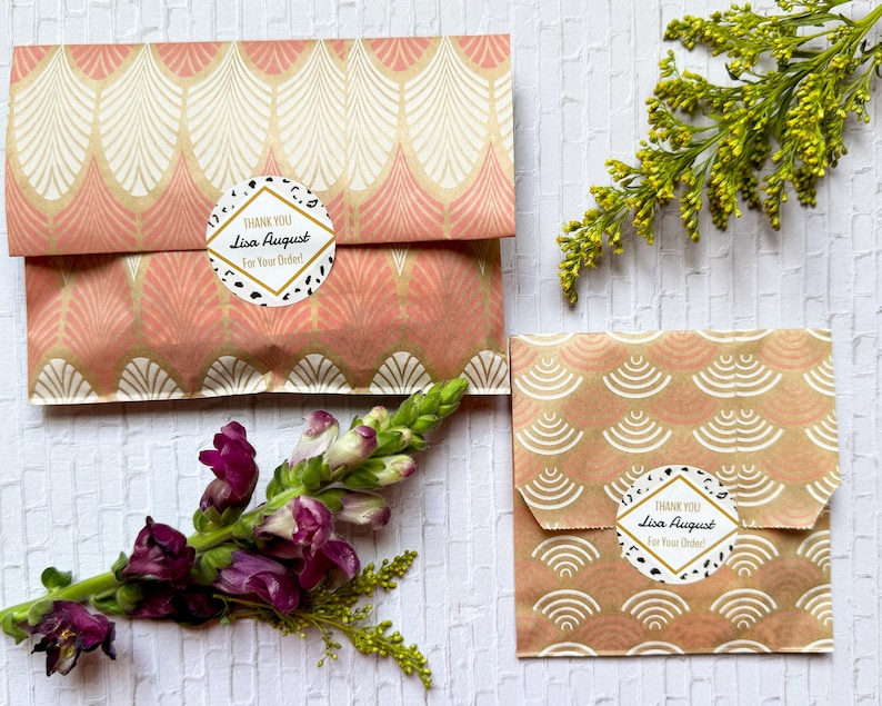 May include: Two small gift bags with a pink and gold geometric pattern. Each bag has a round sticker with black and white polka dots and the text "THANK YOU Lisa August For Your Order!"