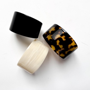 May include: Three wide, rectangular bangles in a top-down view. One is solid black, another is a tortoiseshell pattern of brown and yellow, and the third is off-white with a marbled effect. The bangles are likely made of resin or a similar material.