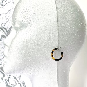 May include: A pair of brown and white tortoiseshell hoop earrings with silver posts. The earrings are on a white mannequin head.