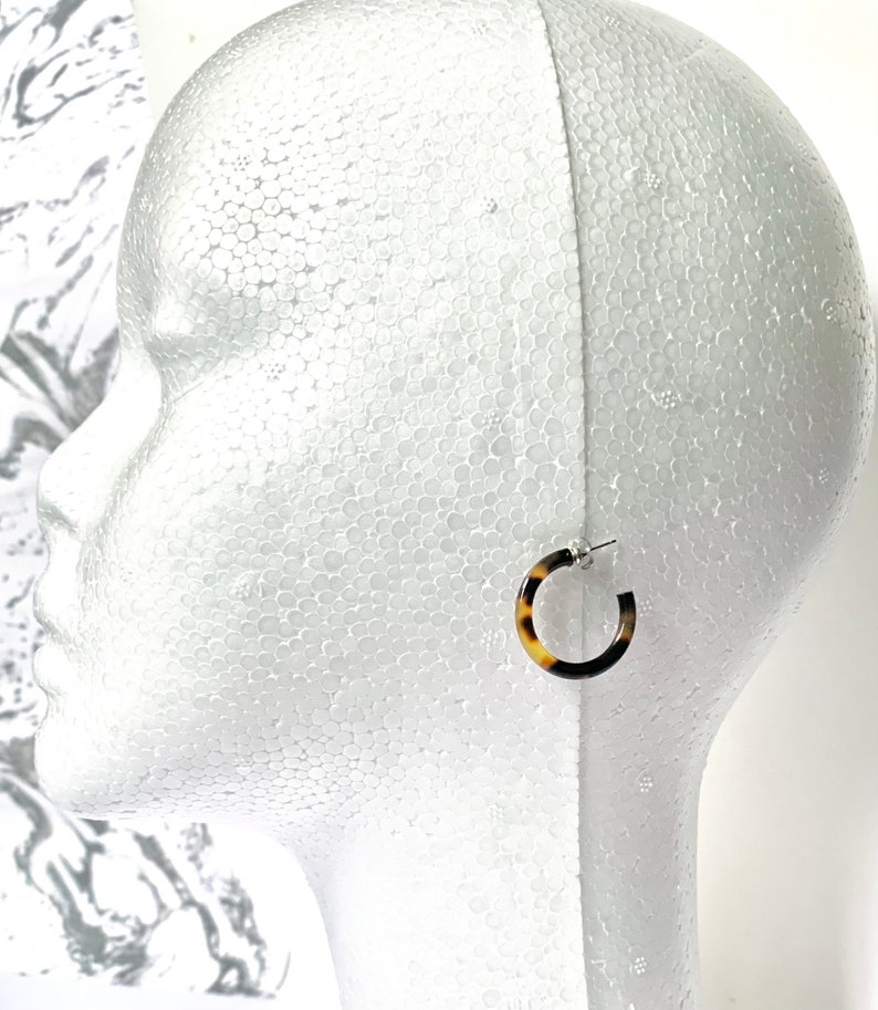 May include: A pair of brown and white tortoiseshell hoop earrings with silver posts. The earrings are on a white mannequin head.