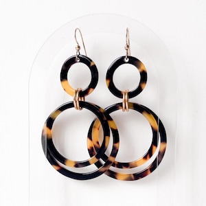 May include: A pair of dangle earrings with a tortoiseshell pattern. Each earring features two connected hoops, one smaller and one larger, in shades of black, brown, and tan. The earrings have gold-colored ear wires and connectors.