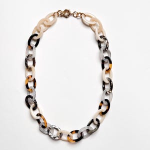 May include: A chunky chain link necklace featuring alternating oval links in cream, translucent gray, and tortoiseshell patterns. The necklace is secured with a gold-toned metal circular spring ring clasp at the top, connecting the two ends of the chain.