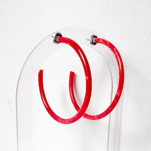 May include: A pair of bright red hoop earrings with a subtle marbled texture. These circular earrings feature a smooth, glossy finish and are secured with silver-toned metal post backings, providing a classic and bold look for any jewelry collection.
