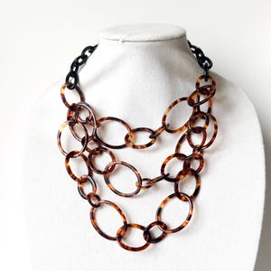 May include: A layered necklace with large oval links in a tortoiseshell pattern. The necklace has a black chain at the back and is displayed on a white mannequin. The necklace is a statement piece.
