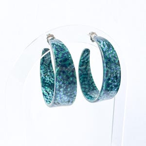 May include: A pair of hoop earrings with a mosaic pattern in shades of teal and blue. The earrings are displayed on a clear stand against a white background. The earrings have silver posts.