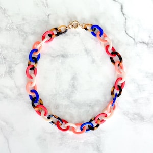 May include: A chunky chain link necklace featuring alternating oval links in solid bright blue, vibrant coral, soft pale pink, and brown tortoiseshell patterns. The necklace is secured with a gold-toned metal circular spring ring clasp.