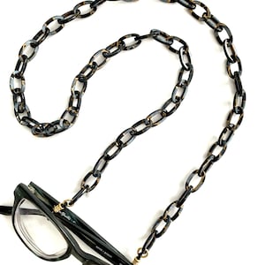 May include: A black and gold chain eyeglass holder with oval links. The chain is attached to a pair of green and black eyeglasses. The eyeglasses have a gold-colored metal detail at the temples.