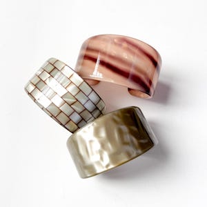 May include: Four wide, open cuff bracelets in varying colors and textures. One is a mosaic of small, rectangular, off-white tiles. Another is a translucent, swirled pink and burgundy. The third is a textured, pearlescent gold. The fourth is a translucent, swirled pink and burgundy.