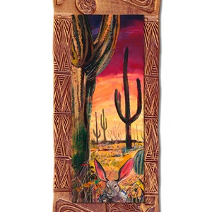 Puede incluir: Una pintura enmarcada que representa una escena del desierto con un conejo asomándose desde detrás de un cactus. El cielo es de un naranja y morado vibrantes, y el cactus es verde con una base marrón. El marco es de madera marrón oscura con tallas intrincadas.