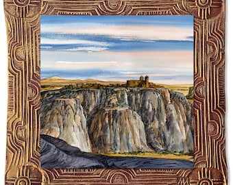 May include: A framed watercolour painting depicting a pueblo village nestled in a canyon. The painting features a blue sky with white clouds, brown cliffs, and a small village with white buildings. The frame is gold with a red border and intricate designs.