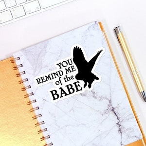 May include: A white keyboard, a gold notebook with a marble cover, a gold pen, a sticker with the text "You remind me of the babe" and a black silhouette of a bird, a pink and yellow macaron on a white plate, a white candle in a glass jar, and two silver paper clips on a white surface.