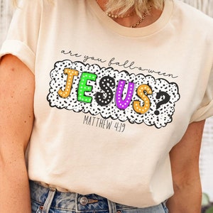 May include: A light beige t-shirt with a black, white, green, and purple graphic that says "Are you Halloween Jesus? Matthew 4:19".