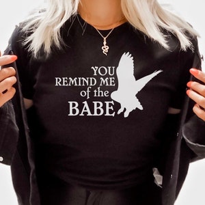 May include: Black crew neck t-shirt with the text "YOU REMIND ME of the BABE" and a white graphic of a bird. The shirt is paired with black leather pants and a black denim jacket. Accessories include a gold necklace and rings.