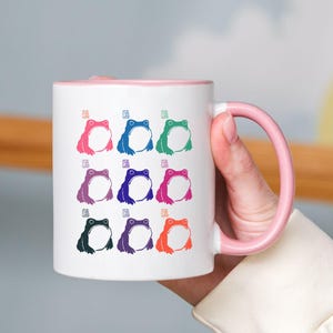 May include: White ceramic mug with a pink rim and handle. The mug features a design of nine frogs in different colors, including blue, green, purple, pink, and orange.