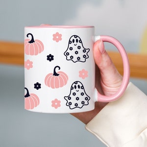 May include: A white ceramic mug with a pink rim and handle. The mug features a repeating pattern of pink pumpkins and black ghosts with white polka dots.