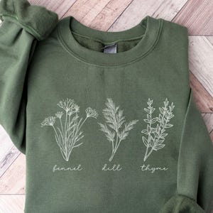 May include: An olive green sweatshirt featuring white botanical illustrations of fennel, dill, and thyme. The names of the herbs are printed below each illustration. The sweatshirt has long sleeves with ribbed cuffs.