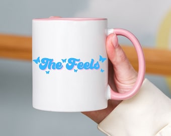 Pode incluir: Caneca de cerâmica branca com borda e alça rosa. A caneca tem um gráfico azul que diz "The Feels" com detalhes de borboletas azuis.