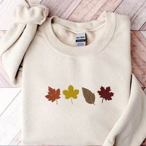 May include: A cream colored sweatshirt with a row of four autumn leaves in shades of red, orange, yellow, and brown. The leaves are embroidered on the front of the sweatshirt.