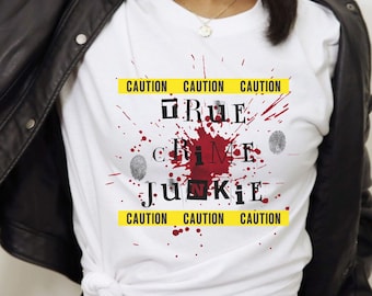 Può includere: Maglietta bianca con un design grafico che dice "TRUE CRIME JUNKIE" in testo nero con schizzi di sangue rosso e impronte digitali. Il testo è circondato da un nastro giallo di attenzione.