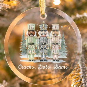 May include: A clear, round Christmas ornament featuring three nutcracker figures in festive uniforms. The ornament has a gold ribbon for hanging and the text "Cracks, Dots, Bams" at the bottom. The background is a blurred image of a Christmas tree.