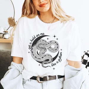 May include: White t-shirt with a black and white graphic of a snake wrapped around a crescent moon. The text "got a hold on me like a Python" is printed on the shirt.