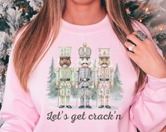 May include: Pink sweatshirt featuring a graphic of three nutcracker soldiers with the text "Let's get crack'n". The design includes a winter scene with evergreen trees. A red mug with a mustache design is also visible.