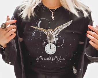 May include: Black t-shirt with a white owl flying through a starry night sky with bubbles. The owl is perched on a clock face that reads 11:59. The text "as the world falls down" is printed below the clock.