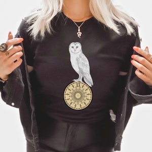 May include: Black t-shirt with a white barn owl perched on a gold circular design with the number "13" in the center. The design features a repeating pattern of crosses and circles.