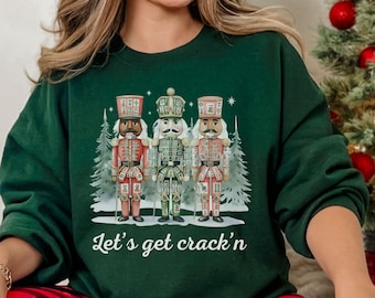 May include: A dark green sweatshirt featuring a graphic of three nutcrackers in red, green, and gold, with a snowy forest backdrop. The text "Let's get crack'n" is printed below the image. The person is wearing red and black plaid pajama pants.