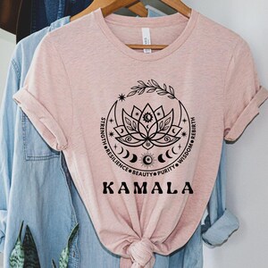 May include: A pink t-shirt with a black graphic of a lotus flower with a crescent moon and stars. The text "STRENGTH, RESILIENCE, BEAUTY, PURITY, WISDOM, REBIRTH" surrounds the flower. The text "KAMALA" is below the graphic.
