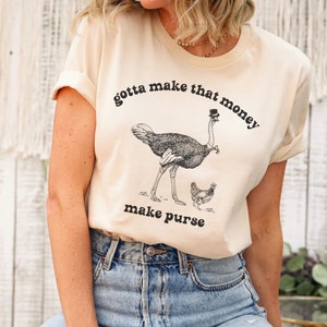 May include: A beige t-shirt with a black and white illustration of an ostrich wearing a top hat and a chicken. The text "gotta make that money make purse" is printed on the shirt.