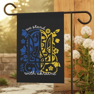 May include: A black garden flag with a yellow and blue Ukrainian trident symbol surrounded by floral designs. The text "we stand with ukraine" is printed below the symbol.