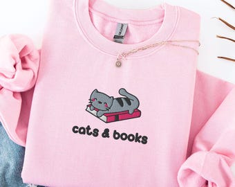 May include: A light pink sweatshirt with a graphic of a gray cat lying on a red book. The text "cats & books" is below the graphic. A gold necklace with a small pendant is on the sweatshirt. Blue jeans are visible in the background.