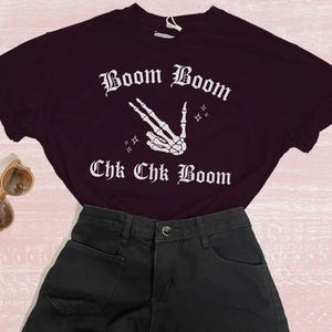 May include: A black t-shirt with white text that reads 'Boom Boom Chk Chk Boom' and a white skeleton hand making a peace sign. The t-shirt is paired with black denim shorts.