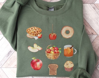 May include: A green sweatshirt with a fall-themed design featuring an apple pie, apples, a donut, a cup of cider, a loaf of bread, and a muffin.