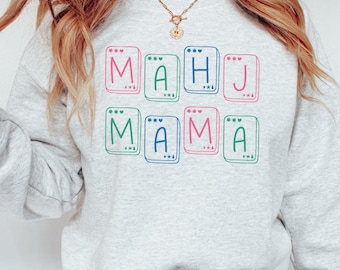 May include: A light gray sweatshirt with the words "MAHJ MAMA" printed in colorful tiles. The letters are in pink, green, blue, and red. The sweatshirt is a casual style.