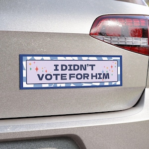 May include: A blue and white bumper sticker with a pink background and colorful stars. The sticker reads "I didn't vote for him."