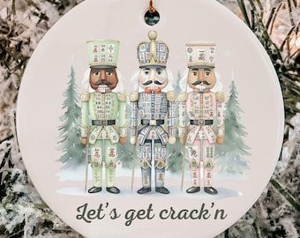 May include: Round Christmas ornament featuring three nutcracker soldiers in green, blue, and pink uniforms. The ornament has the text "Let's get crack'n" and a gold string for hanging. The background shows a snowy scene with pine trees.