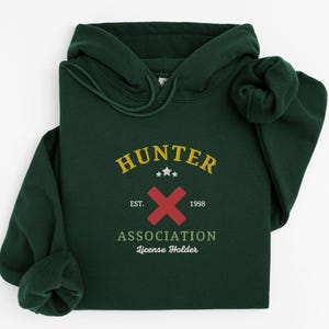 May include: A dark green hoodie with the text "HUNTER" in yellow, a red "X" and the words "EST. 1998" and "ASSOCIATION License Holder". The hoodie has a drawstring and a front pocket.