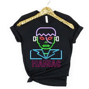 May include: Black t-shirt featuring a neon-style graphic of Frankenstein's monster. The design includes green, red, purple, and blue outlines, with the word "MANIAC" in pink below the image. The shirt is hanging on a gold sequined hanger.