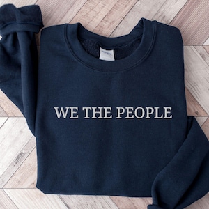 May include: A navy blue crewneck sweatshirt featuring the text WE THE PEOPLE embroidered in white across the center chest. The garment has long sleeves with ribbed cuffs and a ribbed neckline, showcasing a clean and simple design.